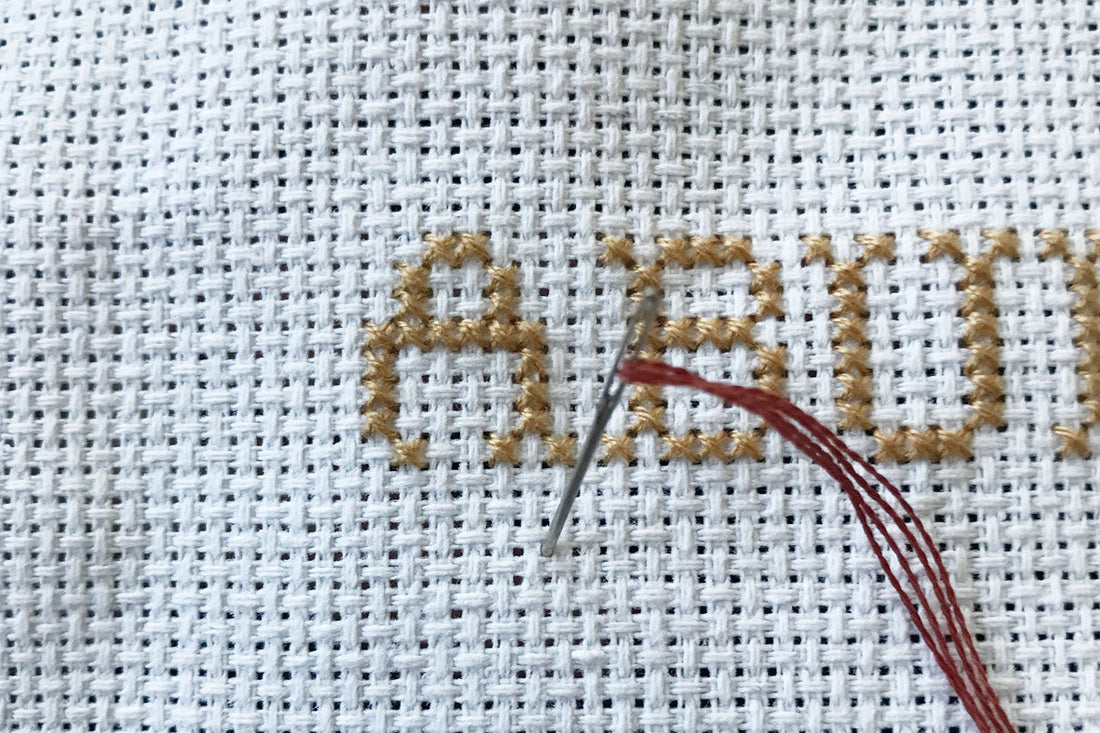 An image showing a needle and thread going through aida fabric to start stitching